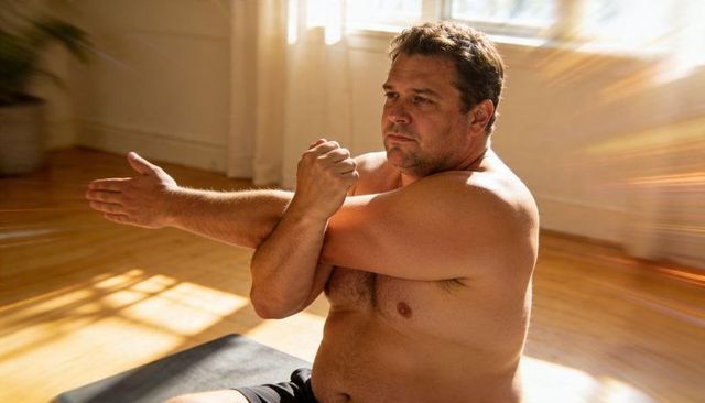 Middle-aged man stretching shirtless on yoga mat in sunlit room promoting wellness and calm