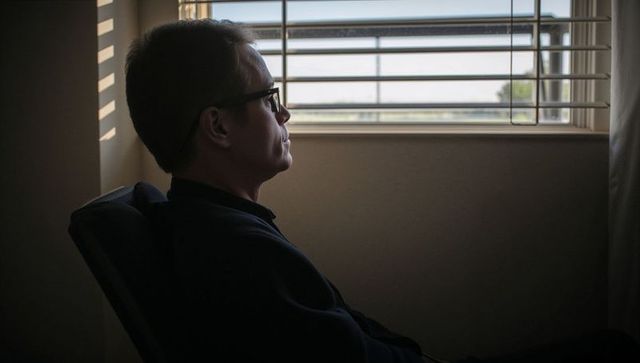 Contemplative man wearing glasses sitting by window blinds, interior portrait moody tone