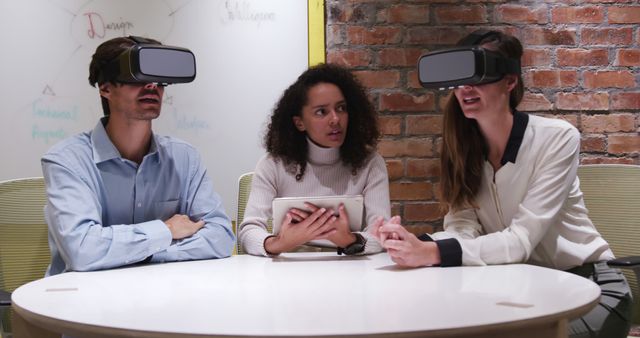 Three coworkers are engaging in a modern meeting utilizing virtual reality technology. Two of them are wearing VR headsets, while a third person holds a tablet and looks interested in the experience. The setting includes a brick wall and a whiteboard, indicating a tech-savvy work environment. This image can be used for presentations, articles, and marketing materials related to technology in the workplace, team collaboration, and innovative business practices.