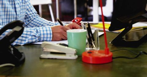 Man writing at office desk with stapler coffee mug and red lamp, workspace productivity