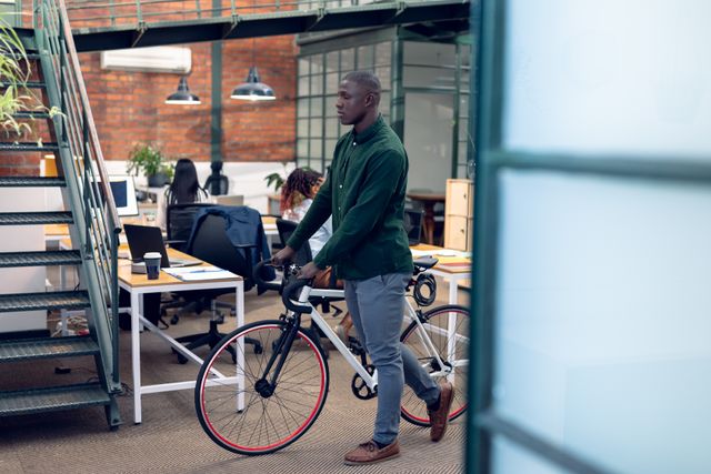 Young African American businessman walking with a bicycle in a modern, creative office environment. Ideal for illustrating themes of sustainable transportation, modern work culture, and urban professional lifestyle. Suitable for use in articles, blogs, and marketing materials related to business, coworking spaces, and eco-friendly commuting.