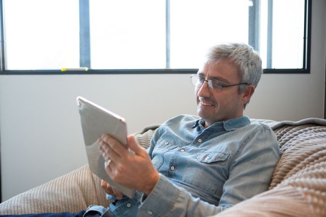 Side of a happy senior Caucasian man working in a creative office, sitting on a armchair, using a digital tablet, smiling