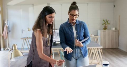 Two female coworkers collaborating over laptop in modern minimalist daylit office workspace