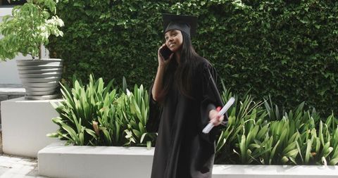 Asian graduate celebrating outdoors holding diploma calling on smartphone