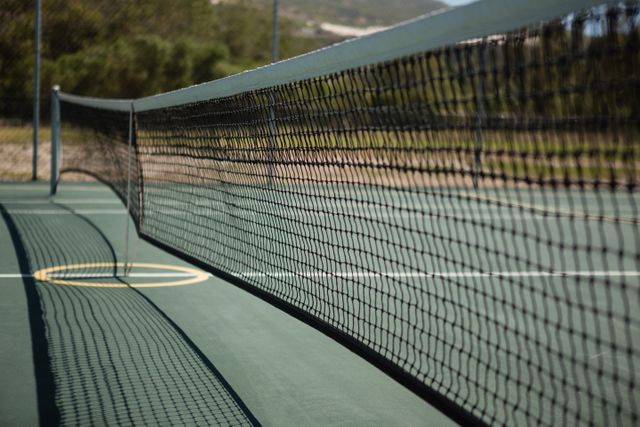 Close-up view of a tennis court net on a sunny day, casting shadows on the green court surface. Ideal for use in sports-related content, fitness promotions, outdoor activity advertisements, and recreational facility brochures.