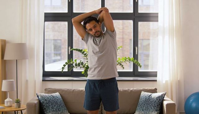 Man stretching in sunlit living room by window, performing morning home exercise routine