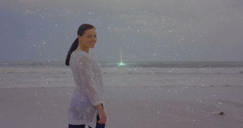 Smiling woman standing on overcast beach wearing sheer white blouse and dark pants, green flare