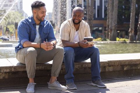 Two friends sitting and chatting over takeaway coffee by city fountain, urban lifestyle