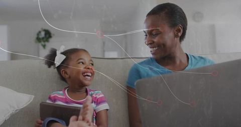 Black mother and daughter laughing on couch using tablet and laptop in cozy home