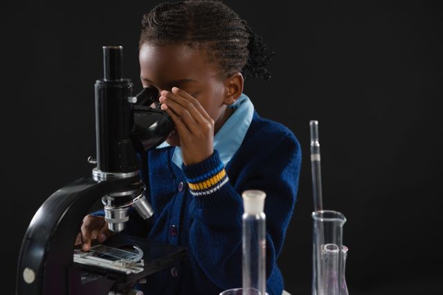 Young schoolgirl in uniform using a microscope, focusing on a slide. Laboratory glassware is visible on the table. Ideal for educational materials, science class promotions, STEM programs, and children's learning resources.