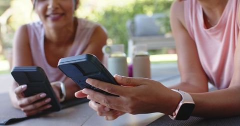 Multiracial women relaxing on yoga mats checking smartphones and smartwatch on sunlit patio