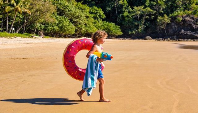 Child walking on sunlit tropical beach with pink donut float, towel and water blaster