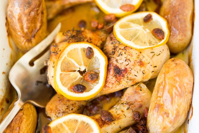 Close-up of a delicious roasted chicken with lemon slices and whole baked potatoes in a baking dish. The dish is garnished with herbs and a savory brown sauce. Ideal for illustrating recipes, food blogs, family dinner ideas, catering services, restaurant menus, and food magazines.
