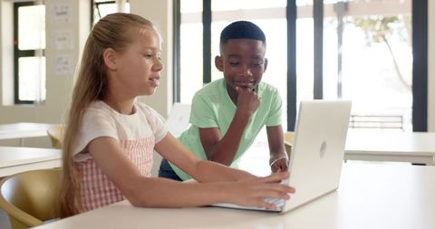 Diverse students collaborating on laptop in bright classroom, teamwork and learning