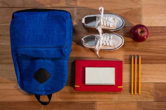 This image shows a neatly arranged collection of school essentials on a wooden table. Items include a blue backpack, white sneakers, a red notebook, three pencils, and a red apple. Ideal for use in educational materials, back-to-school promotions, and organizational tips for students.