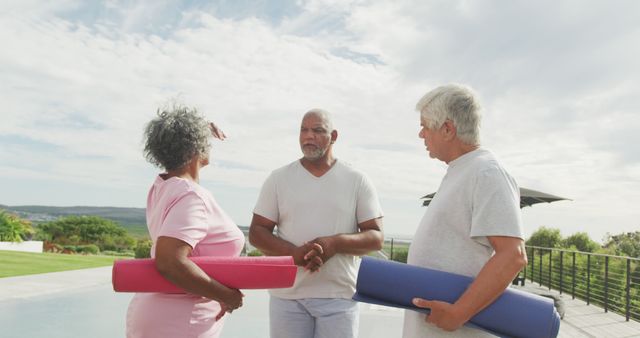 Three senior individuals standing outdoors, each holding a yoga mat, suggesting they are preparing for a yoga or fitness session. A serene natural background indicates a peaceful, health-focused activity setting. Suitable for promoting senior fitness, wellness retreats, and healthy outdoor lifestyles.