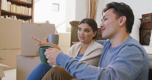 Couple is seated on floor surrounded by moving boxes, enjoying coffee and taking a break from unpacking. Ideal for illustrating themes of moving, new beginnings, home lifestyle, and relationships.