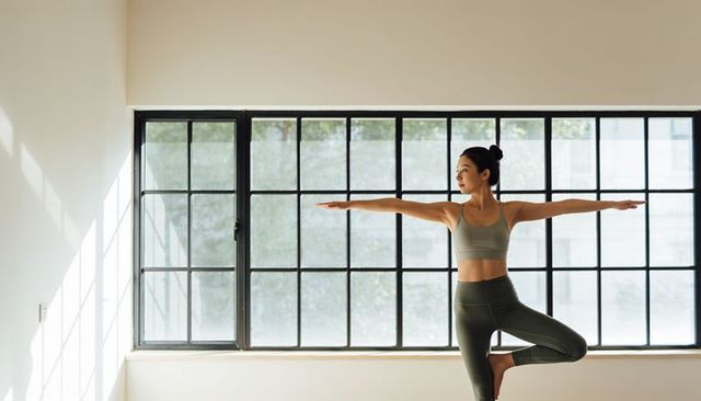 Asian woman practicing yoga in minimalist studio by large black window with natural light