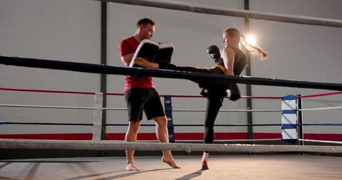 Female boxer delivering high roundhouse kick while coach holding pads in boxing ring
