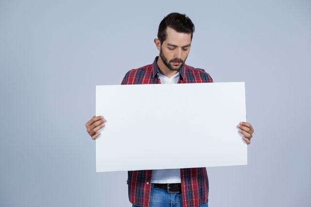 Young man in casual attire holding a blank placard against a grey background. Ideal for advertisements, messages, or announcements. Perfect for use in marketing materials, social media posts, or promotional content where customizable text or graphics are needed.