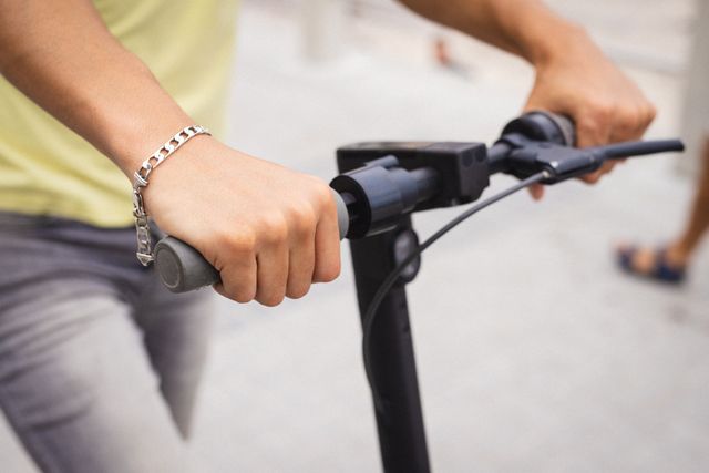 This image shows a close-up of a young man's hands holding the handlebar of a push scooter. The man is wearing a bracelet, and the background is slightly blurred, indicating an urban setting. This image can be used for topics related to personal transportation, urban mobility, outdoor activities, and modern lifestyle.