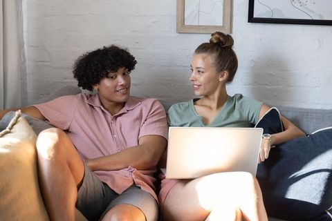 Young couple relaxing on couch with laptop, sharing smile and chat in sunlit living room