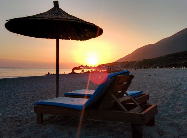 Pairs of lounge chairs under an umbrella on a serene beach at sunset with a beautiful horizon. Ideal for serene vacation advertisements, travel brochures, and websites promoting beach destinations and romantic getaway packages.