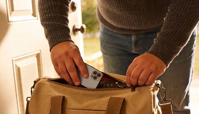Man packing smartphone and key fob into canvas duffel bag at sunlit front door for travel