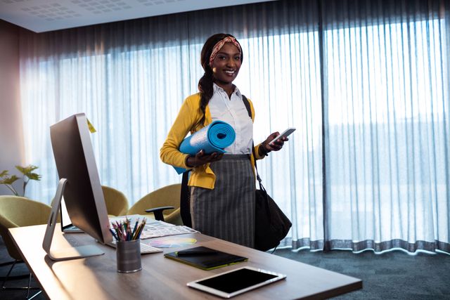 African-American businesswoman holding yoga mat and smartphone in modern office. Incorporates wellness into daily routine, promoting work-life balance. Ideal for themes of workplace wellness, productivity, and healthy living in a corporate environment.