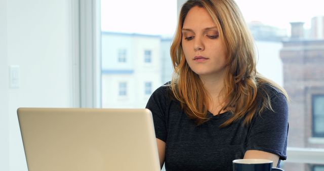 Young woman working on her laptop at home. She appears to be focused and productive. Suitable for themes relating to remote work, freelancing, technology, business, and modern work environments.