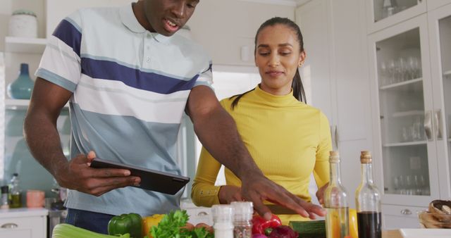 This image portrays a happy diverse couple preparing food together in a modern kitchen while using a recipe on a tablet. It encapsulates themes of domestic life, teamwork, and inclusivity, making it suitable for promotions about healthy living, family bonding, and modern lifestyle marketing. Brands focused on kitchen appliances, cooking classes, or inclusive family products could utilize this image effectively.