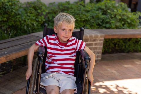 Young blond boy sitting in wheelchair on sunlit brick courtyard wearing red striped shirt