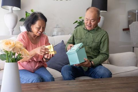 Asian senior couple exchanging gifts and smiling on cozy sofa at home with pastel decor