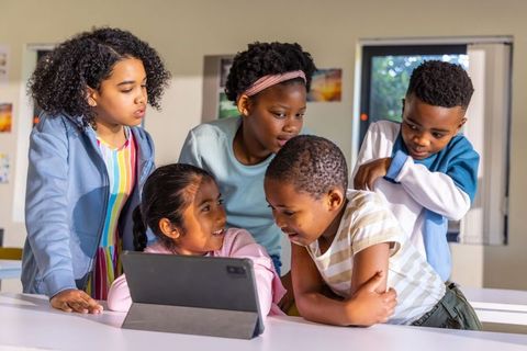 Diverse children gathering around tablet in classroom collaborating on digital learning