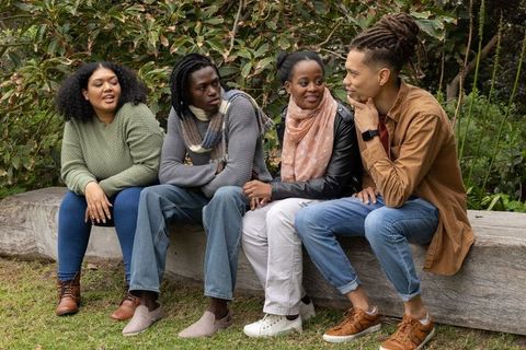 Friends chatting on wooden log bench in leafy park wearing scarves and casual outfits