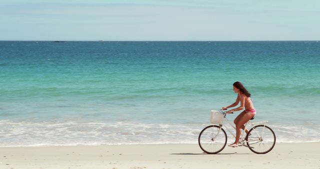 Woman riding bicycle on sandy beach with turquoise ocean waves in the background, enjoying a sunny day. Ideal for concepts related to outdoor activities, summer holidays, relaxation, beach adventures, and healthy lifestyles.