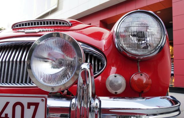 Close-up of the front view of a classic red vintage car highlighting the headlights and bumper. The detailed shot showcases the car's design and craftsmanship, perfect for use in automotive publications, vintage car enthusiast blogs, classic car restoration businesses, or for illustrating historical advancements in car designs.