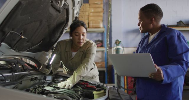 Image of two diverse female car mechanics talking, using laptop and repairing car. working in car repair shop and running small feminine business concept.