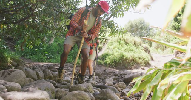 Man is traversing a rocky path through a lush forested area, utilizing hiking poles for stability. Ideal for use in content about outdoor activities, physical fitness, hiking gear, and nature exploration. Could be featured in travel blogs, adventure magazines, and fitness websites.