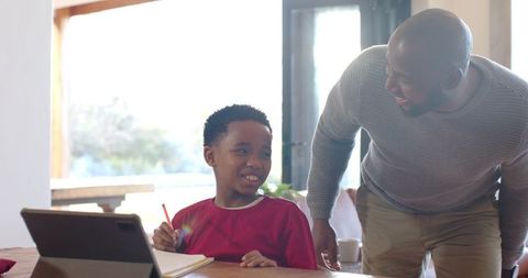 African american father helping son with homework using tablet at bright home dining table