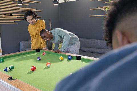 Leaning over pool table, diverse friends studying billiard shot in modern lounge for casual game