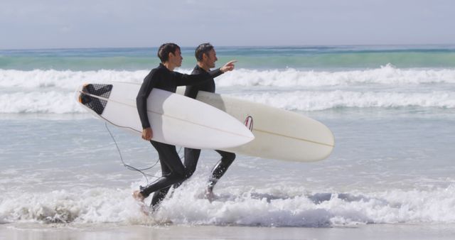 Two men in wetsuits carrying surfboards walking towards the waves. They seem to be discussing or pointing towards the horizon, suggesting excitement or anticipation. This photo can be used to depict themes like beach activity, sports, surfing lifestyle, friendship, and outdoor adventure.
