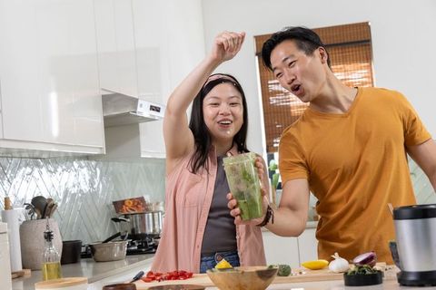 Diverse couple preparing green smoothie in bright home kitchen, holding blender jar