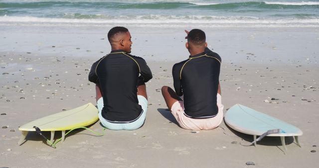 Two young men are sitting on the beach with surfboards next to them, discussing the waves. This image is ideal for promoting beach activities, surfing lessons, summer vacation destinations, or friendship and leisure time spent outdoors. It captures the relaxed atmosphere of beach life and the connection between friends.