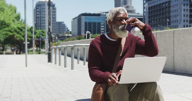 Senior man with gray beard using laptop and drinking coffee in an urban environment. Modern and dynamic lifestyle captured outdoors in a city. Suitable for imagery related to freelance work, remote jobs, senior citizens' tech-savvy lifestyles, urban living, relaxation, and cityscape dynamics.