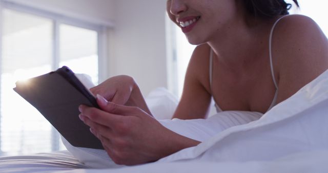 Woman lying in bed and using a tablet for reading or browsing the internet. This image can be used to symbolise relaxation, the convenience of technology, or a cozy home environment. It's ideal for articles or advertisements about home leisure activities, modern lifestyle, or ergonomic comfort in daily life.