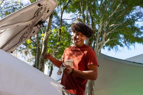 Man checking oil dipstick wiping rag smiling beside open hood car in sunlit driveway
