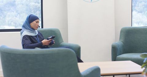 Young woman wearing blue hijab checking smartphone while relaxing in modern teal conference lounge