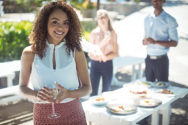 Smiling woman holding a glass of wine at an outdoor restaurant gathering with friends in the background. Ideal for use in lifestyle blogs, social media posts, advertisements for restaurants or wine brands, and articles about social gatherings and leisure activities.