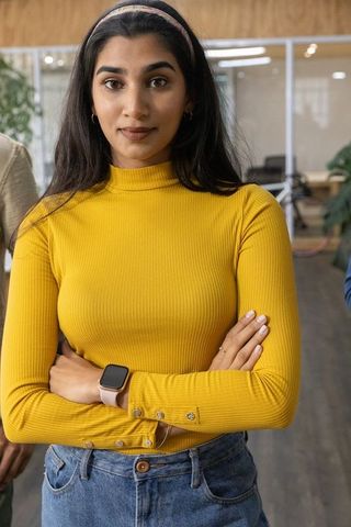 Confident young indian woman standing arms crossed in modern open plan office in mustard top